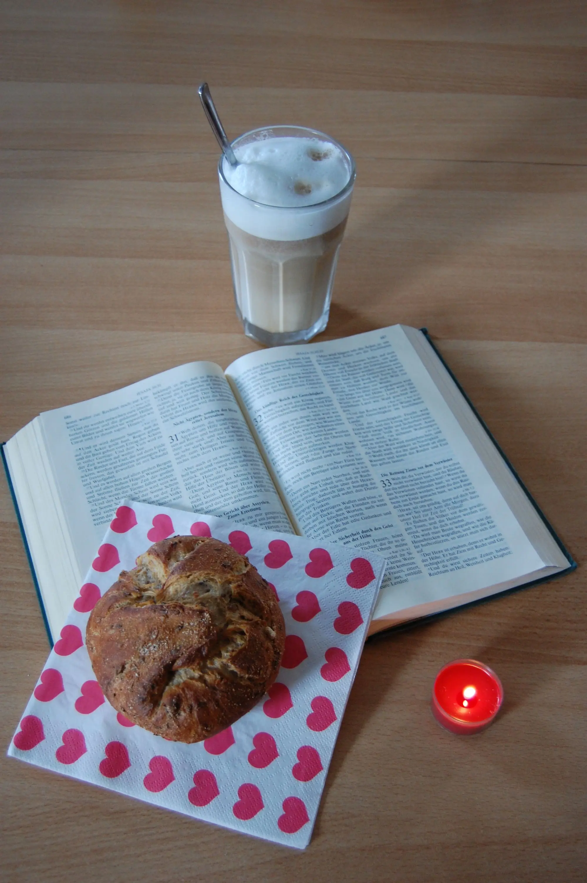 Das Foto zeigt eine aufgeschlagene Bibel mit einer brennenden Kerze, einem Körnerbrötchen und einem Latte Macchiato daneben.