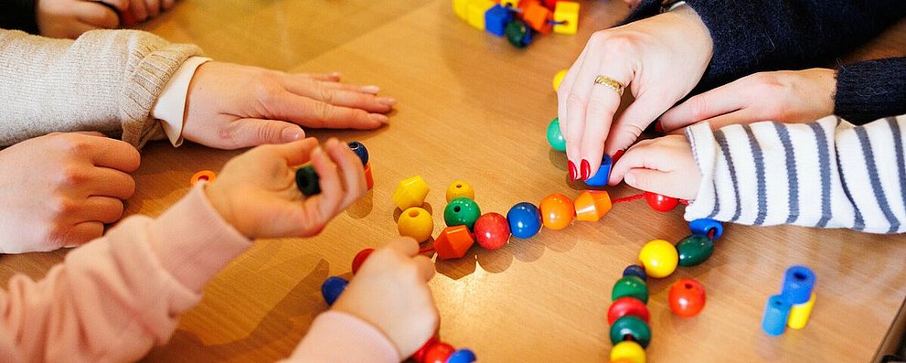 Kita - Kinder beim Spielen. An einem Tisch sitzen einige Kinder, eine Erzieherin und spielen mit einer Kette mit bunten Kugeln und anderen Formen, die auf einer Schnur aufgereiht sind.