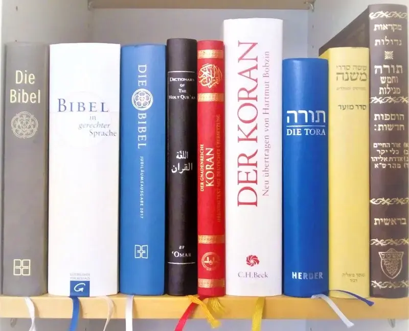 In einen Bücherregal stehen nebeneinander je drei verschiedene Ausgaben der Bibel, des Korans sowie zwei Ausgaben der Tora und eine Mischna