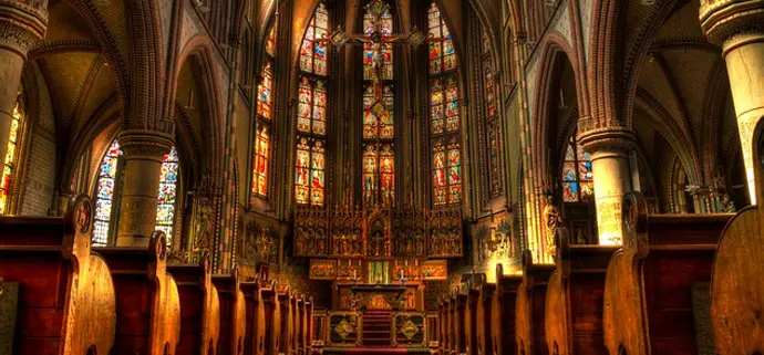 Der Innenraum einer beeindruckenden, reich geschmückten Kirche ist zu sehen. Hohe Gewölbebögen, bunt leuchtende Kirchenfenster und ein prunkvoller Altar dominieren das Bild. Auf beiden Seiten stehen Reihen hölzerner Kirchenbänke.