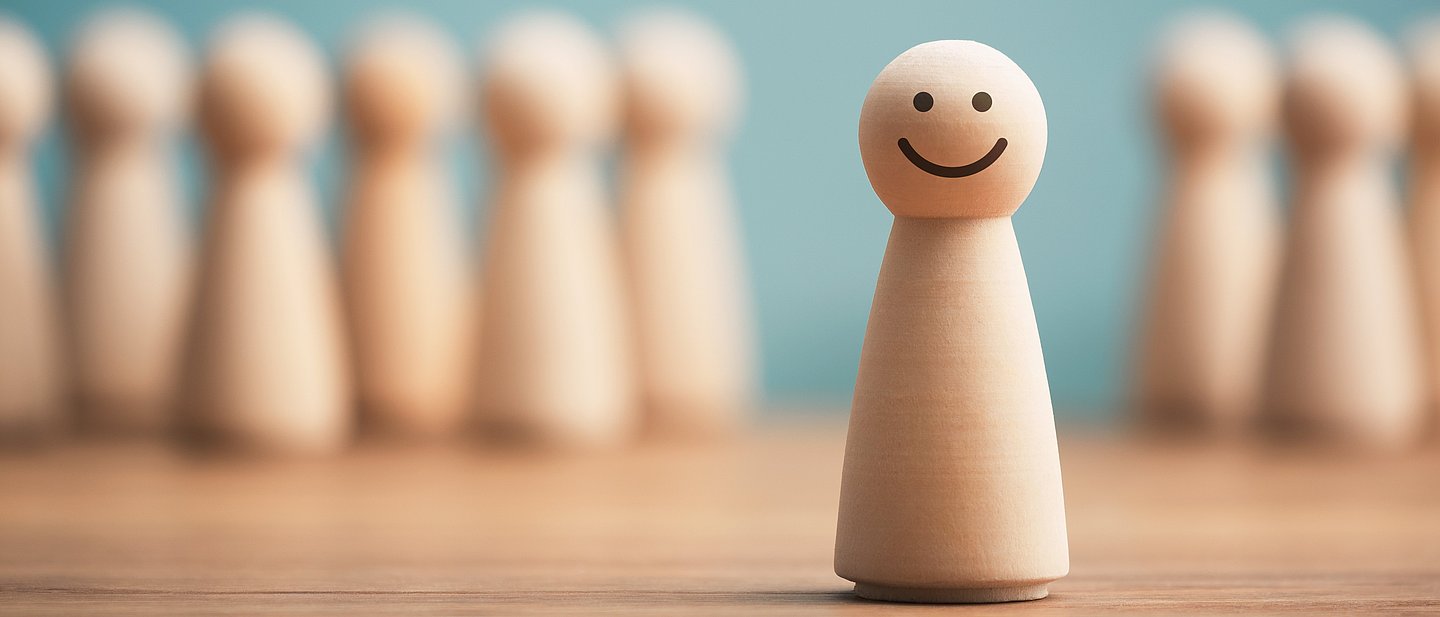 Ein Holzkegelmännchen mit lächelndem Gesicht steht auf einer Tischplatte. Im Hintergrund ist unscharf eine Reihe von neun Holzkegelmännchen zu sehen