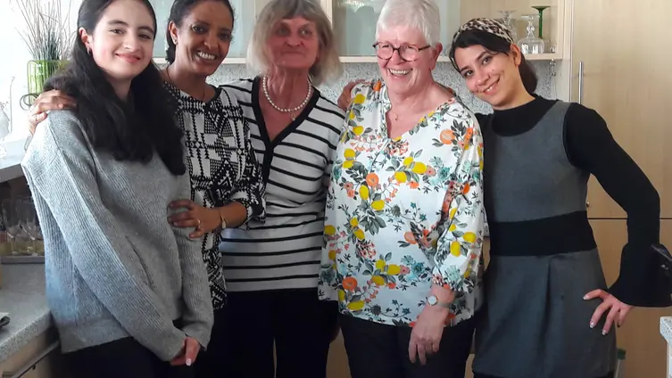 Das Bild zeigt eine Gruppe von fünf Frauen, die nebeneinander stehen und lächeln. Sie scheinen in einem häuslichen Umfeld zu sein, möglicherweise einer Küche, da im Hintergrund Schränke sichtbar sind. Die Frauen haben unterschiedliche Ethnien und Altersgruppen, und ihre Kleidung ist vielfältig. Sie wirken freundlich und entspannt.