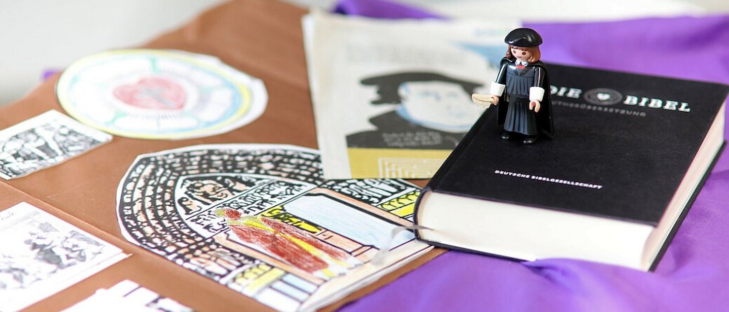 Aufnahme von vorne. Luther trägt schwarzen Talar, Hut und Feder in der Hand