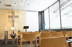 Leerer Andachtsraum mit Holzkreuz an der Wand, Altar und mehreren Stuhlreihen. Große Fenster lassen viel Tageslicht herein und schaffen eine ruhige, helle Atmosphäre.