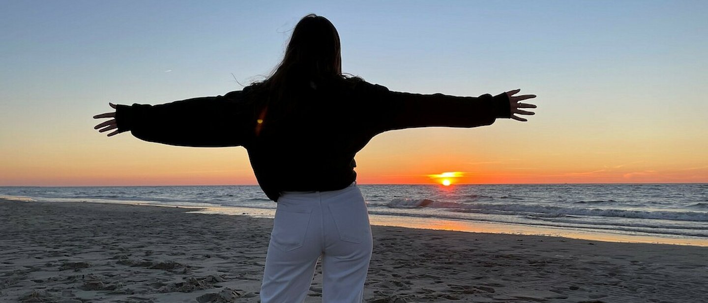 Eine Person steht mit ausgebreiteten Armen am Strand und schaut auf den Sonnenuntergang über dem Meer
