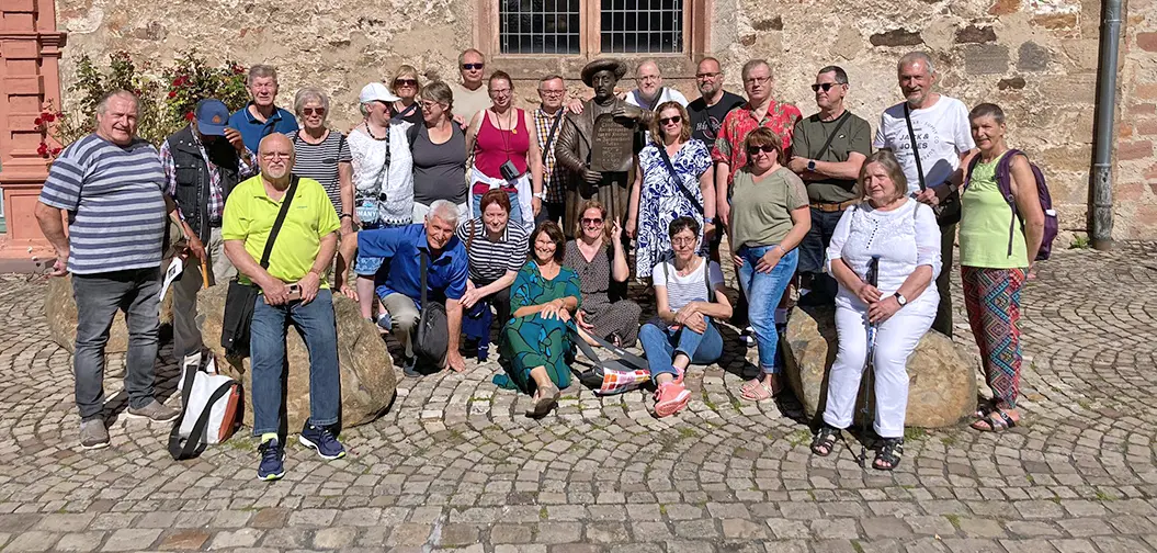 Gruppe von Menschen posiert bei sonnigem Wetter vor einer Steinmauer neben einer bronzenen Statue.