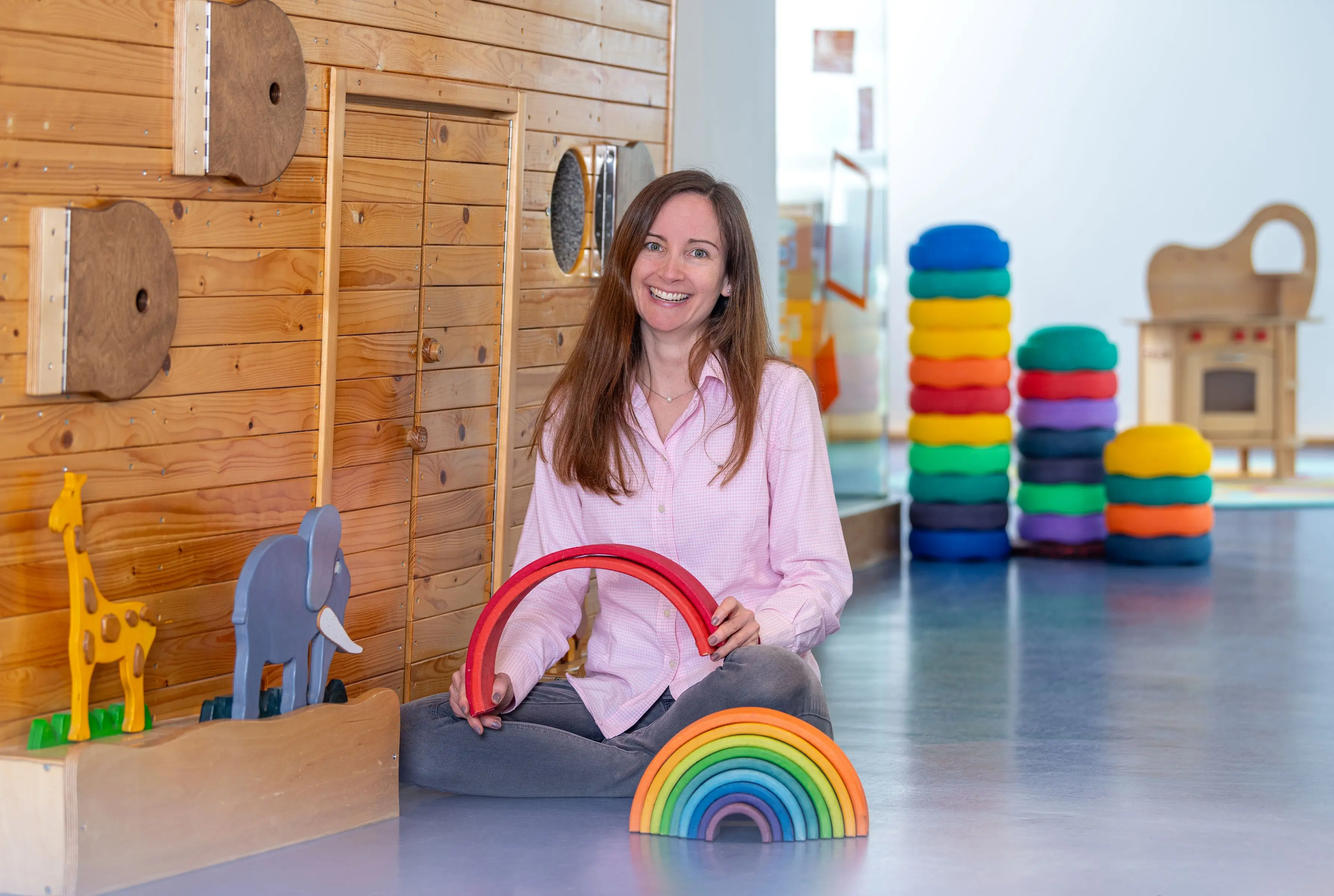 Eine Frau in rosa Bluse sitzt auf dem Boden in einem Kindergarten vor einer Holzwand, die die Arche Noah darstellt. Im Vordergrund ein Regenbogen aus Holzbögen, von denen die Frau zwei in der Hand hält. Am linken Rand eine kleine Holzgiraffe und ein kleiner Holzelefant. Im Hintergrund sind bunte Stapelsteine gestapelt. 