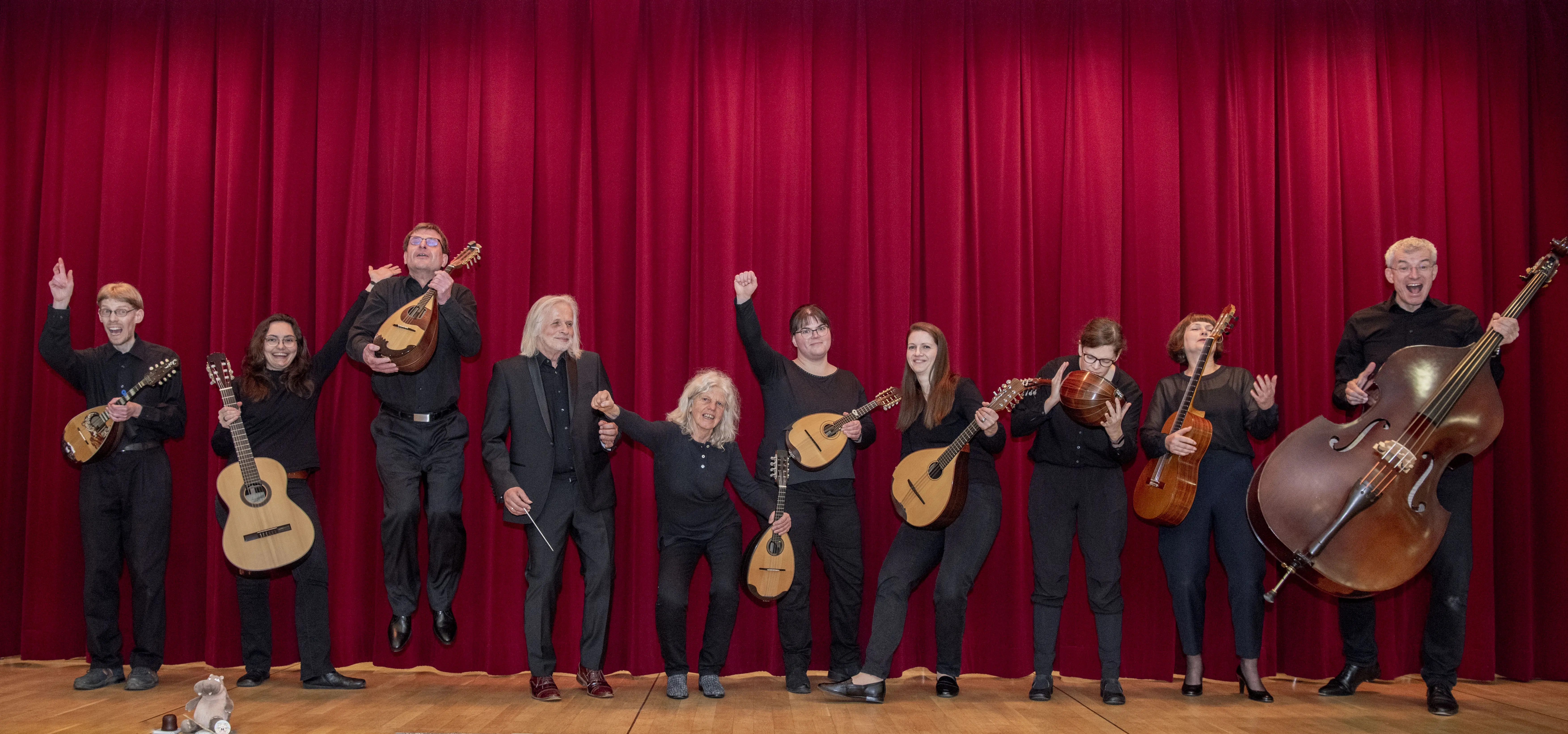 Hier stehen dier Musikerinnen und Musiker des Mandolinen-Orchesters Neuenhain vor einem roten Bühnenvorhang. Alle Musiker:innen sind schwarz gekleidet und haben ihr Instrument in der Hand. Sie lachen, machen Verrenkungen und zeigen damit ihren Spaß am gemeinsamen Musizieren. 