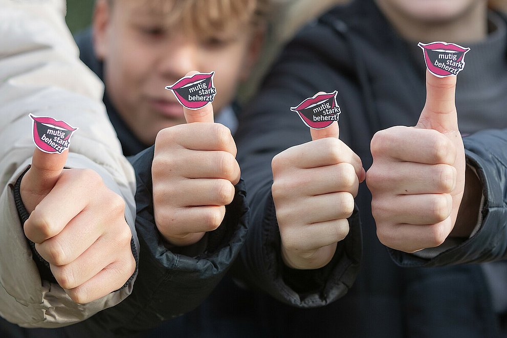 Zwei Konfirmanden und zwei Konfirmandinnen tragen das Logo des Kirchentages 2025 in Hannover (Offener Mund mit den Worten "mutig", "stark" und "beherzt") an ihrem Daumen.