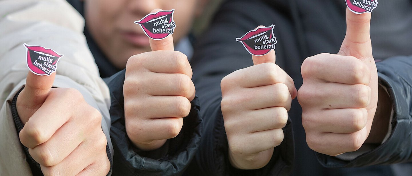 Zwei Konfirmanden und zwei Konfirmandinnen tragen das Logo des Kirchentages 2025 in Hannover (Offener Mund mit den Worten "mutig", "stark" und "beherzt") an ihrem Daumen.