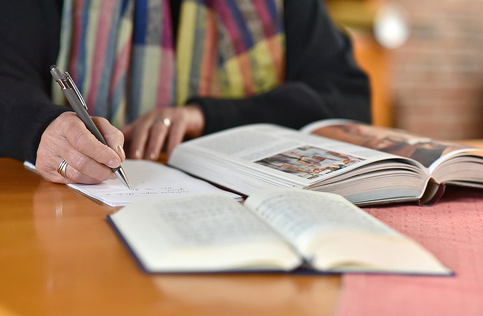 Auf einem Tisch liegen eine Bibel und ein Block. Man sieht zwei Hände, die einen text schreiben.