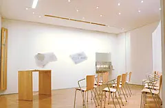 Heller Raum der Stille mit schlichten Holzstühlen, kleinen Tischen und dezenten Kunstobjekten auf Podesten. Die minimalistische Gestaltung und das warme Licht schaffen eine ruhige und meditative Atmosphäre.