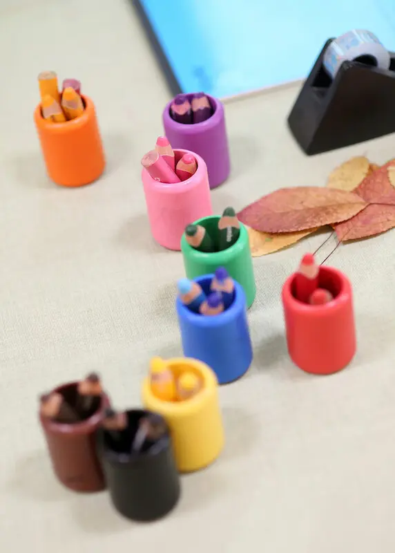 Auf einem Tisch stehen kleine Becher mit kurzen dicken Buntstiften, farblich sortiert, dahinter ein Abroller für Klebeband.