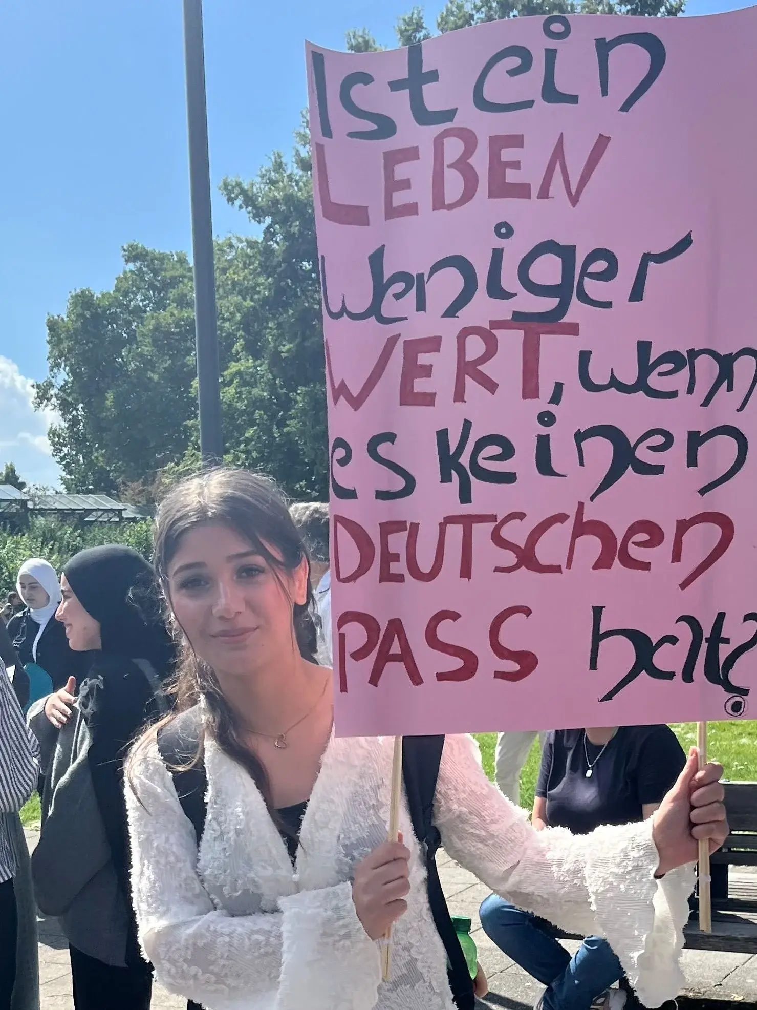 Jugendliche hält ein pinkes Plakat auf dem steht: Ist ein Leben weniger wert, wenn es keinen deutschen Pass hat?