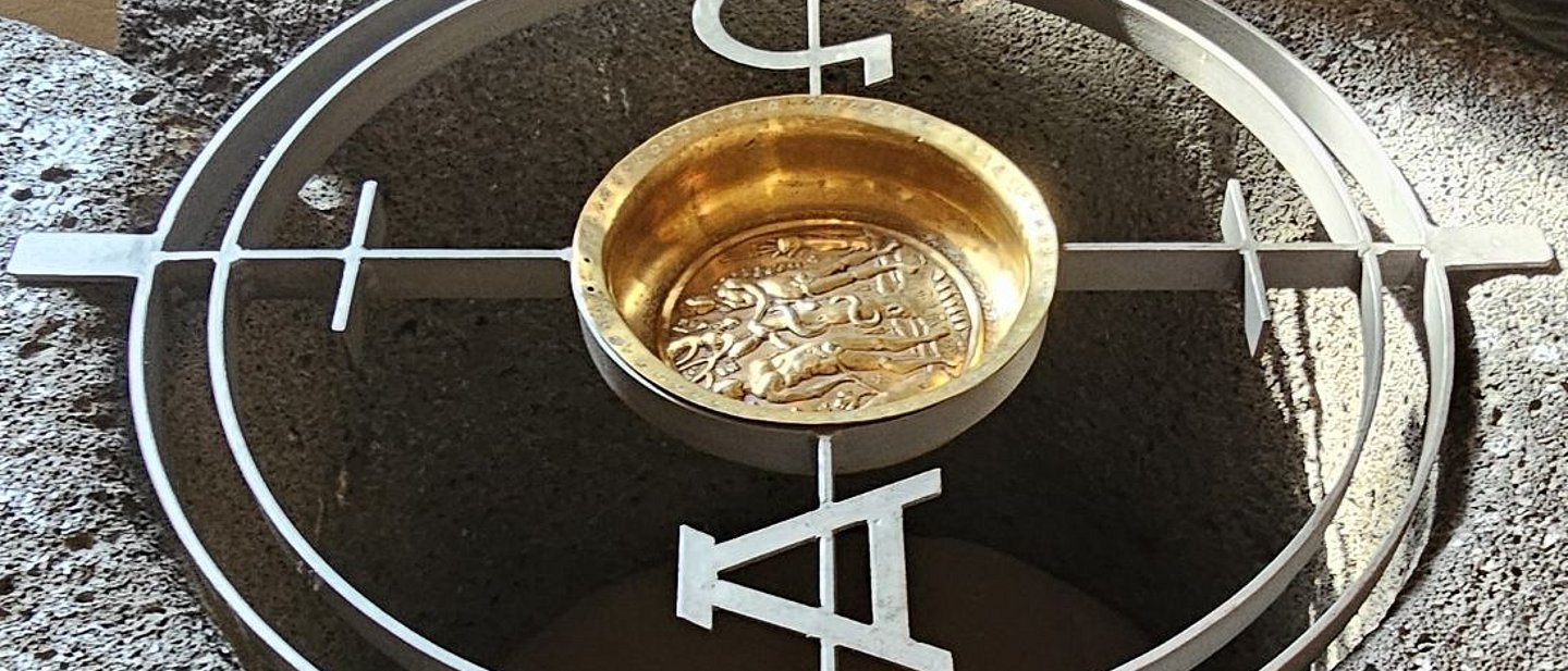 Steinumrandun mit Metallkreis, goldener Taufschale und Alpha und Omega