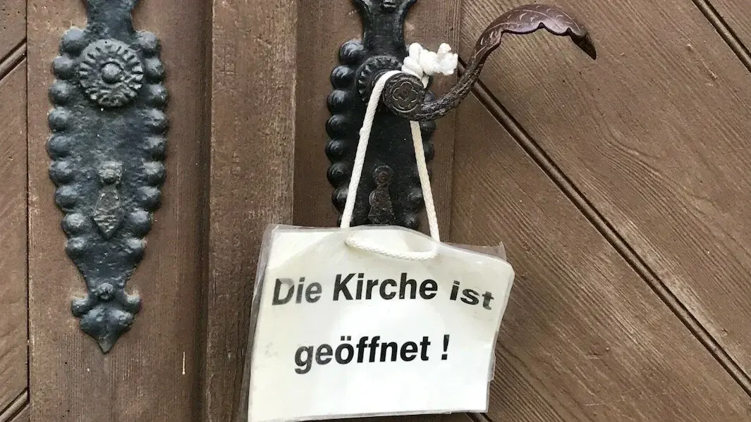 Weißes Schild hängt an Klinke der Kirchentür. Auf dem Schild steht: "Die Kirche ist geöffnet".