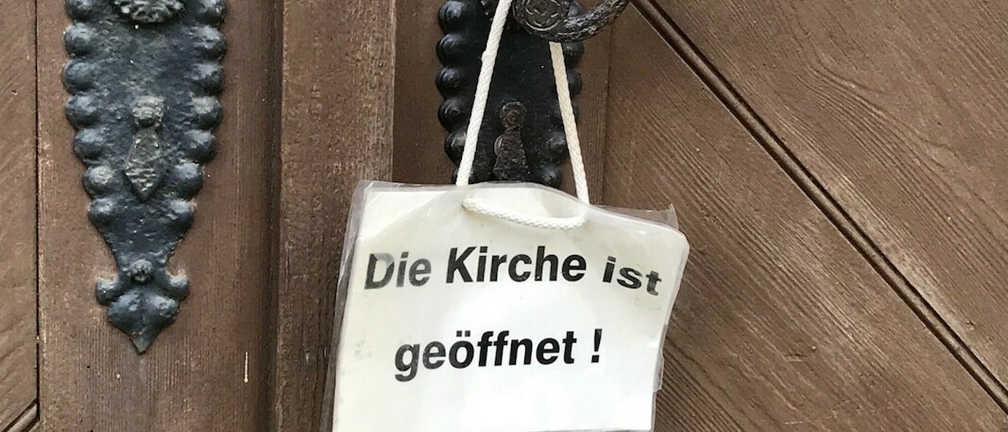 Weißes Schild hängt an Klinke der Kirchentür. Auf dem Schild steht: "Die Kirche ist geöffnet".