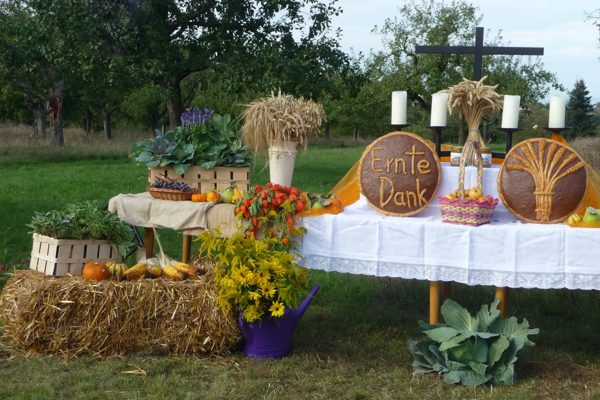 Ein Erntedankaltar, der auf einer Wiese aufgebaut ist. Im Hintergrund sieht man Bäume und Hecken. Der Altar ist mit einer weißen Tischdecke bedeckt. Darauf stehen in Vasen Ähren, Blumen, Obst, Kürbisse und zwei große Brote, außerdem ein schlichtes Kreuz und vier große weiße Kerzen. Vor dem Altar liegt ein Strohballen, darum auch Blumen und Gemüse.