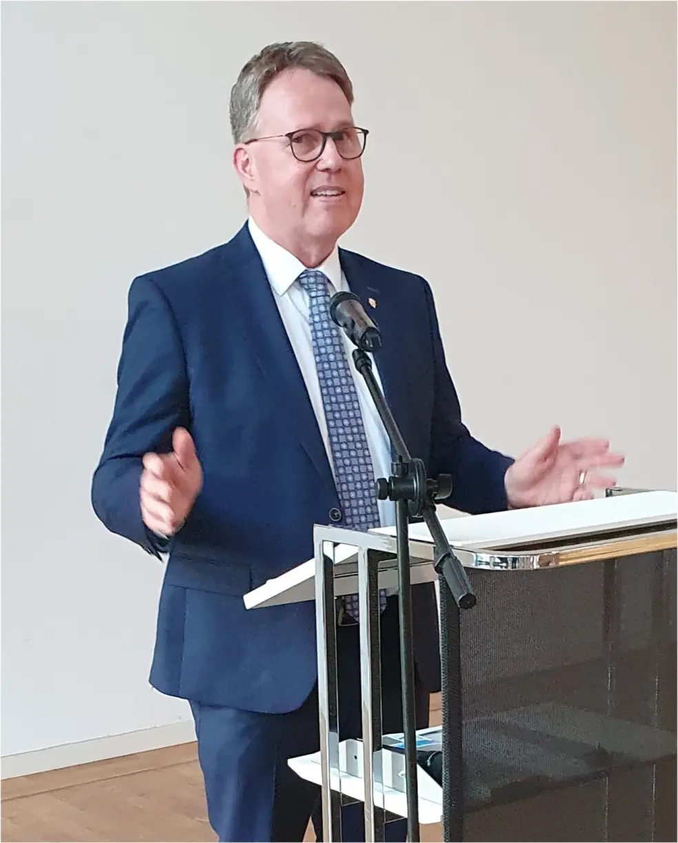 Pfarrer Michael Grimm steht an einem Rednerpult und spricht. Er trägt einen blauen Anzug.