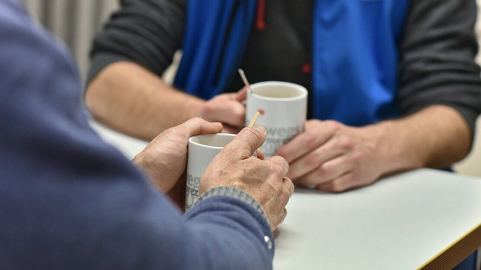 Mitarbeiter der Bahnhofsmission beim Beratungsgespräch. Mitarbeiter und Klient sind ohne Gesichter zu sehen mit Kaffeetasse in den Händen. Fokus auf Hände