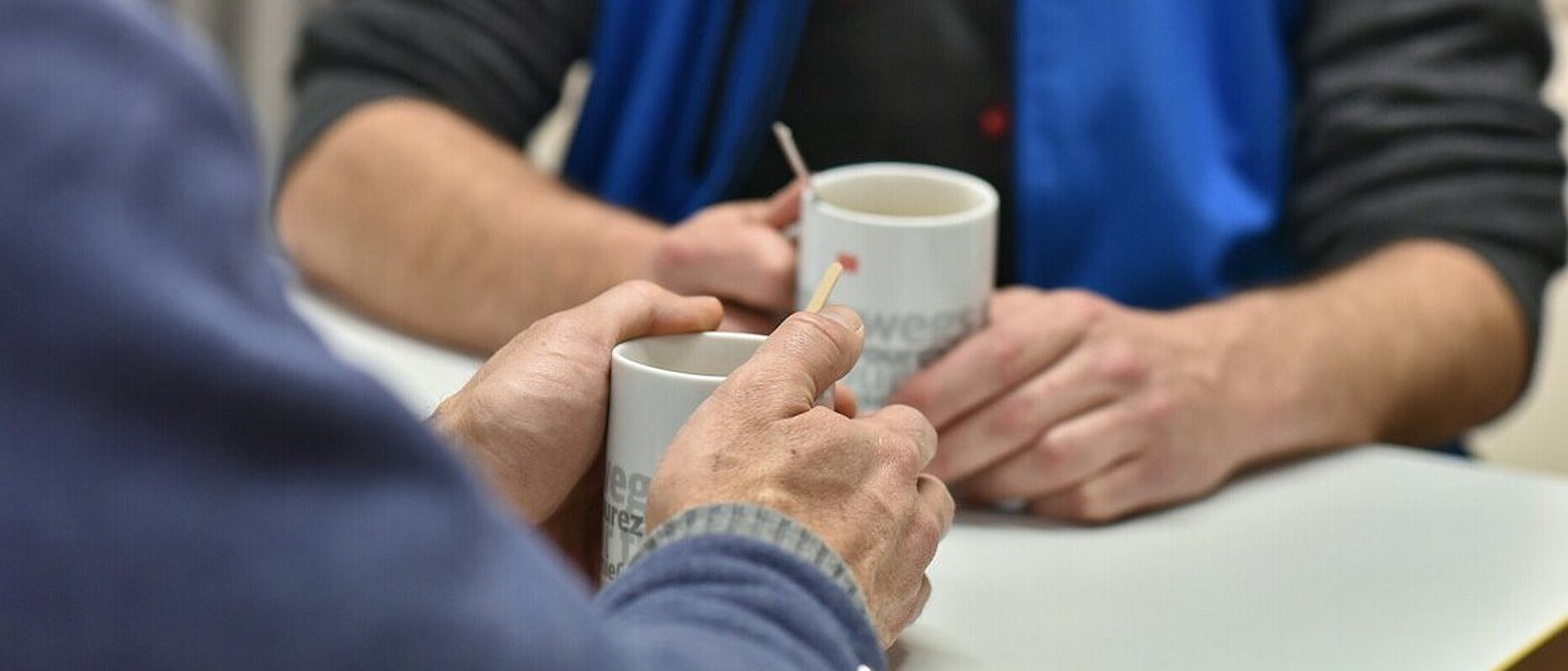 Mitarbeiter der Bahnhofsmission beim Beratungsgespräch. Mitarbeiter und Klient sind ohne Gesichter zu sehen mit Kaffeetasse in den Händen. Fokus auf Hände