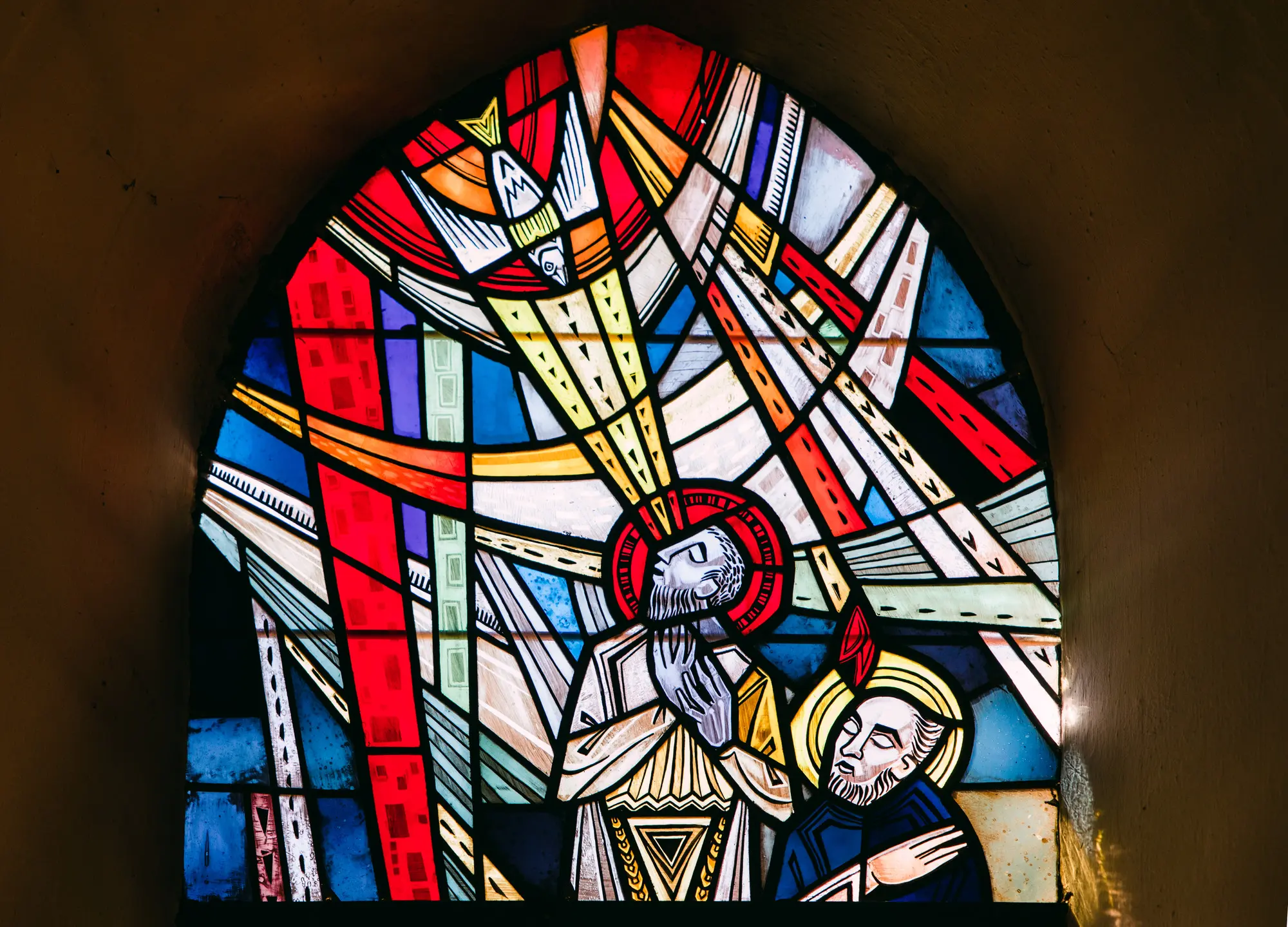 Das Bild zeigt ein großes und bundes Kirchenfenster. Dargestellt ist, wie der Heilige Geist herabfährt auf zwei Menschen.