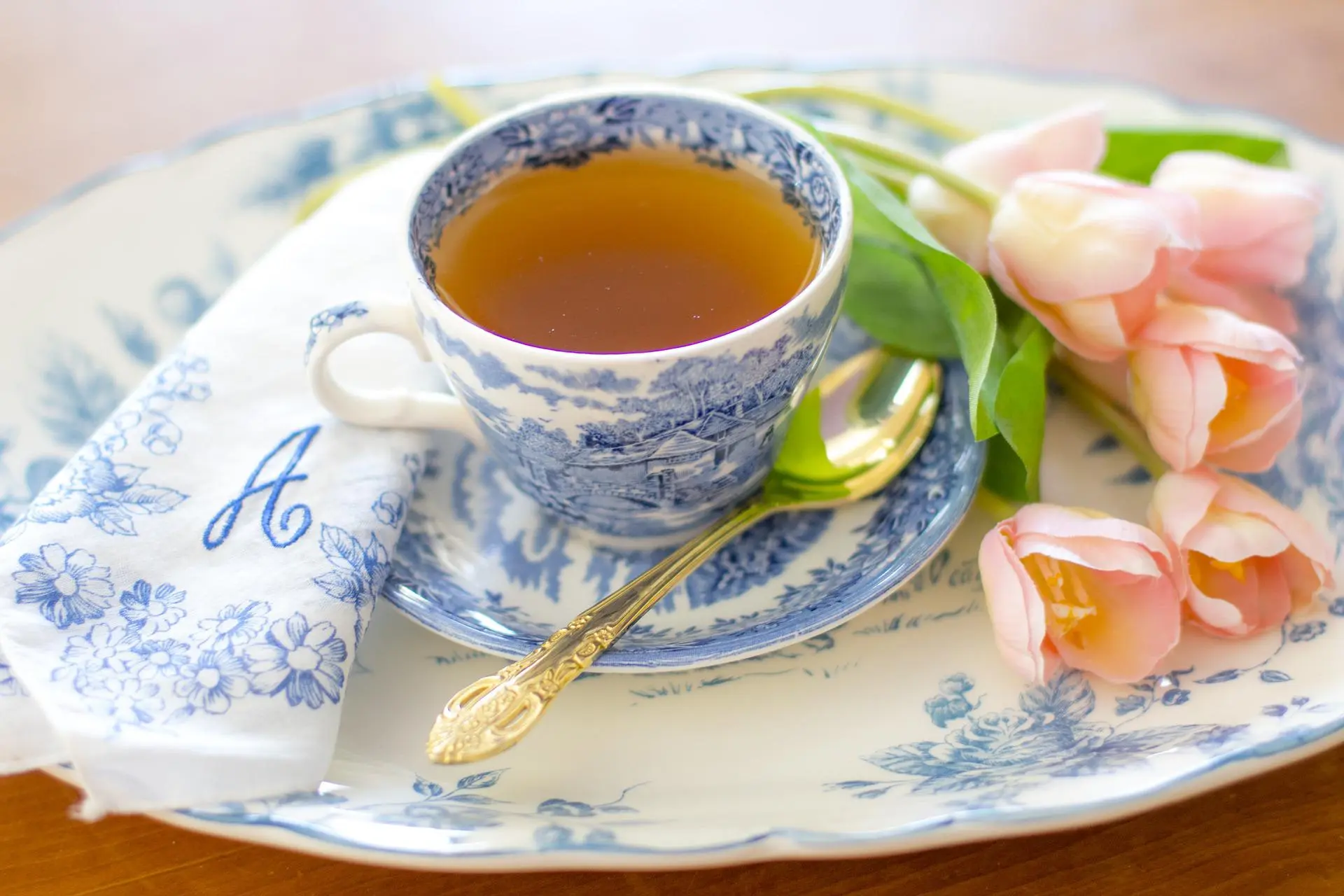 Eine gefüllte Teetasse steht auf einem Kuchenteller, daneben liegen eine Serviette und ein goldfarbener Teelöffel.