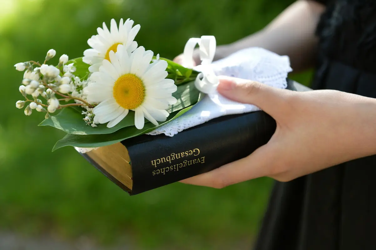 Hände einer Konfirmandin, die Gesanbuch und Blumen halten