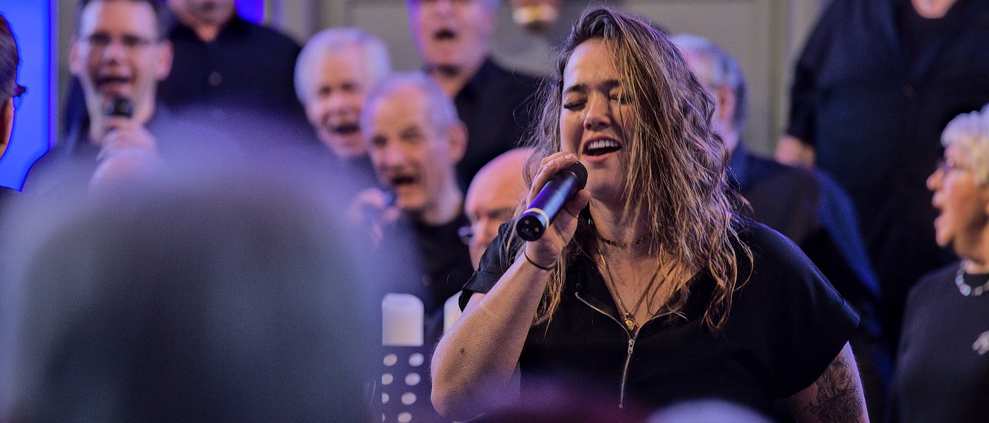 Man sieht aus dem Publikum in Gottesdienstreihen eine blond gelockte Sängerin in Schwarz die als Solistin singt. Im Hintergrund weitere Sänger des Gospelchores Rejoice, auch in schwarz gekleidet.