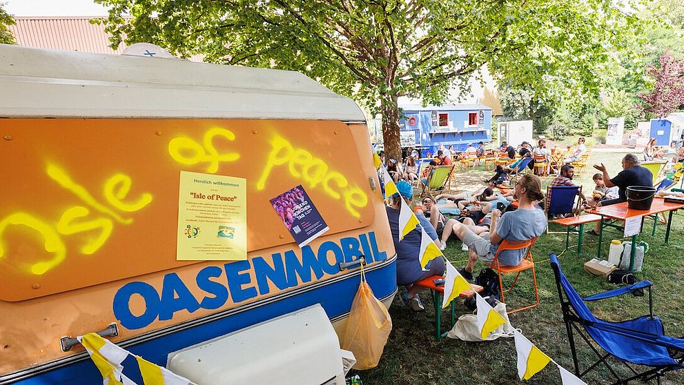 Man sieht im Vordergrund das Oasenmobil und im Hintergrund Jugendliche auf Bänken und Stühlen