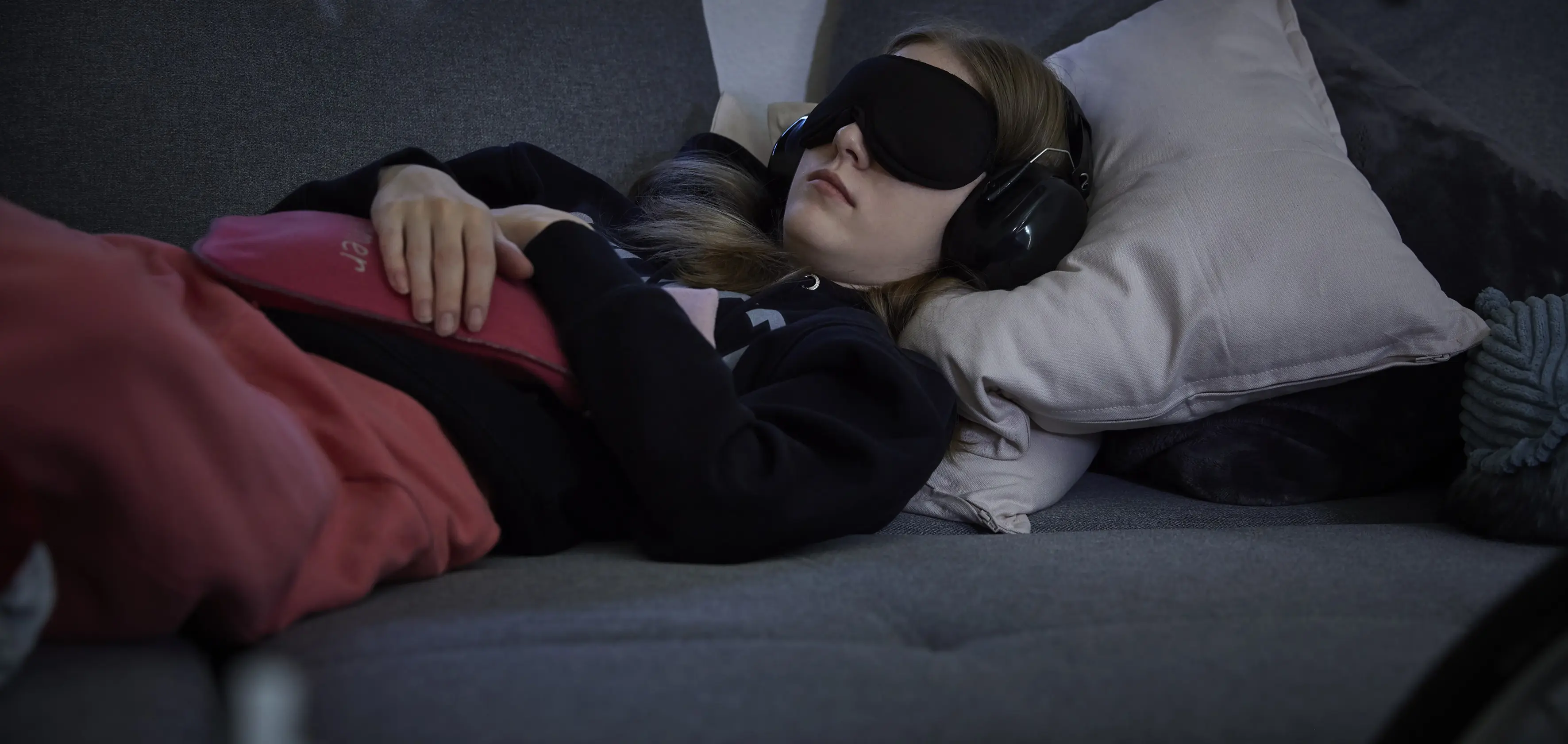 Eine junge Frau liegt auf einem Sofa. In den Händen hält sie eine Wärmflasche mit rotem Bezug. Die Augen sind mit einer schwarzen Schlafbinde bedeckt. Auf den Ohren trägt sie Kopfhörer/Schallschutz.