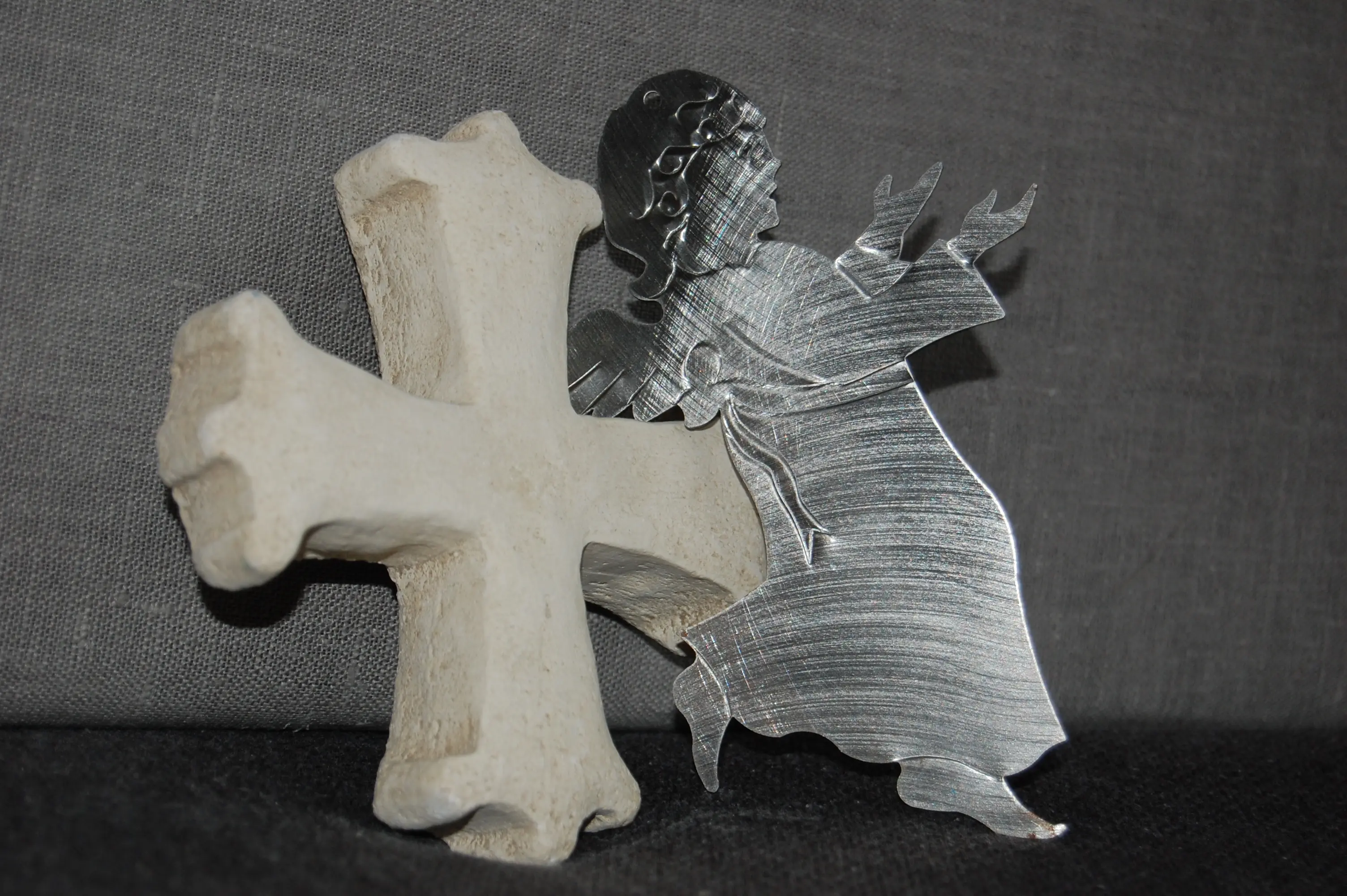 Das Foto zeigt ein helles Kreuz und einen Engel aus silbernem Metall der die Hände zum Himmel geöffnet hat.