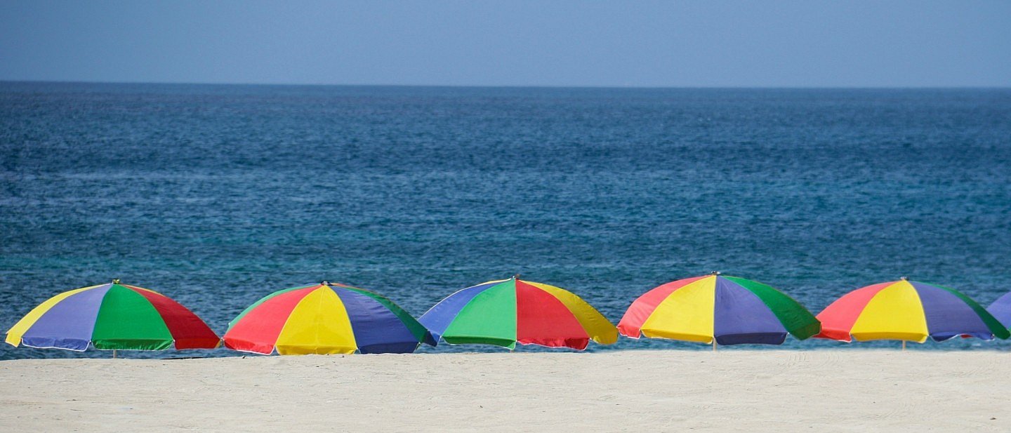 Bunte Sonnenschirme in einer Reihe am Strand, dahinter das Meer