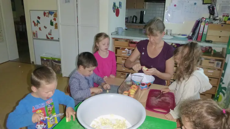 Auf dem Foto sieht man mehrere Kinder, die gemeinsam mit einer Erzieherin Äpfel in eine große Schüssel schneiden.