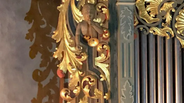Vergoldete und mit Blattwerk verzierte linke Wange des Orgelprospekts in der evangelischen Kirche Köppern. Darin sind einige Pfeifen zu sehen. In dem Blattwerk der Oberkörper eines Kindes/Engels mit Posaune.