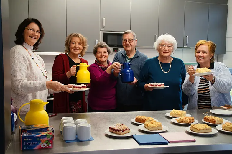 Gruppe älterer Menschen steht in einer Küche, hält Kaffeekannen und Kuchenteller und lächelt in die Kamera.
