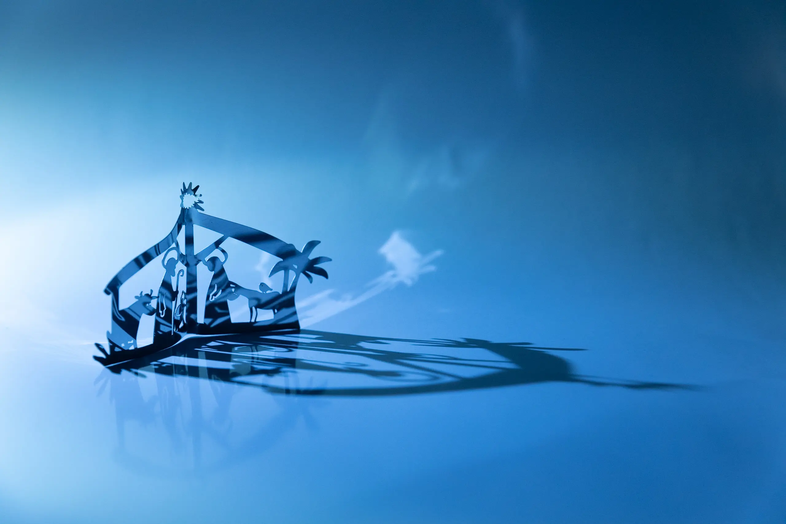 Weihnachtskrippe mit Maria, Josef, Jesus in der Krippe, Ochse und Esel sowie einer Palme, Scherenschnitt, evtl. aus Metall, vor strahlend blauem Hintergrund, spiegelt sich auf der blauen Fläche. Die Krippe nimmt nur einen kleinen Teil des Bildes ein.