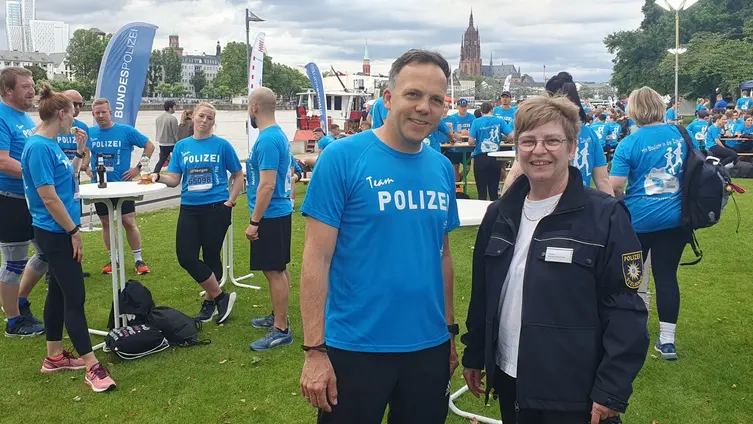 Am  Mainufer stehen viele Läuferinnen und Läufer der Polizei in hellblauen T-Shirts. Im Vordergrund unterhalten sich der verantwortliche Polizeileiter und die Pfarrerin.