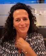 Porträt einer lächelnden Frau mit dunklen lockigen Haaren. Sie trägt eine gemusterte Bluse und sitzt in einem Innenraum mit modernem, unscharfem Hintergrund. Das Bild zeigt Frau Roth.