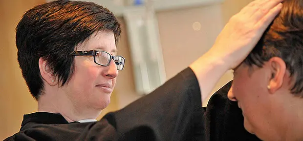 Segnungsszene in einem Kirchenraum. Eine Pfarrerin mit Brille legt einer Frau fürsorglich die Hand auf den Kopf. Beide Personen sind seitlich zu sehen, die Atmosphäre wirkt ruhig und persönlich.
