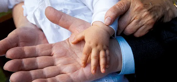 Die Hände von drei Generationen liegen übereinander: Eine kleine Kinderhand ruht auf der geöffneten Hand eines Erwachsenen, während eine ältere Hand die Kinderhand sanft hält. Das Bild vermittelt Nähe, Fürsorge und Verbundenheit.