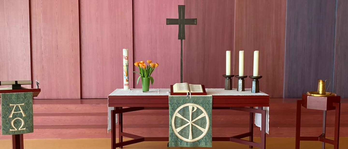 Bild des Altars der Andreasgemeinde, links ist ein Pult zusehen, in der Mitte der Altar mit Kreuz, Kerzen und der Bibel. Rechts daneben steht das Taufbecken