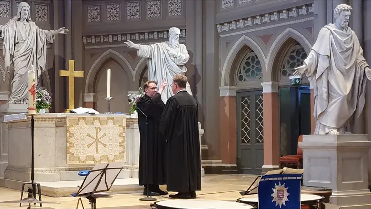 Vor dem Altar der Marktkirche Wiesbaden stehen zwei Pfarrer in schwarzen Talaren. Im Hintergrund große Steinfiguren von Jesus Christus und zwei Aposteln.