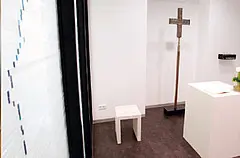 Kleiner heller Andachtsraum mit schlichtem Holzkreuz, weißem Hocker und einem schmalen Altar. Die minimalistische Gestaltung vermittelt Ruhe, Stille und Besinnung.