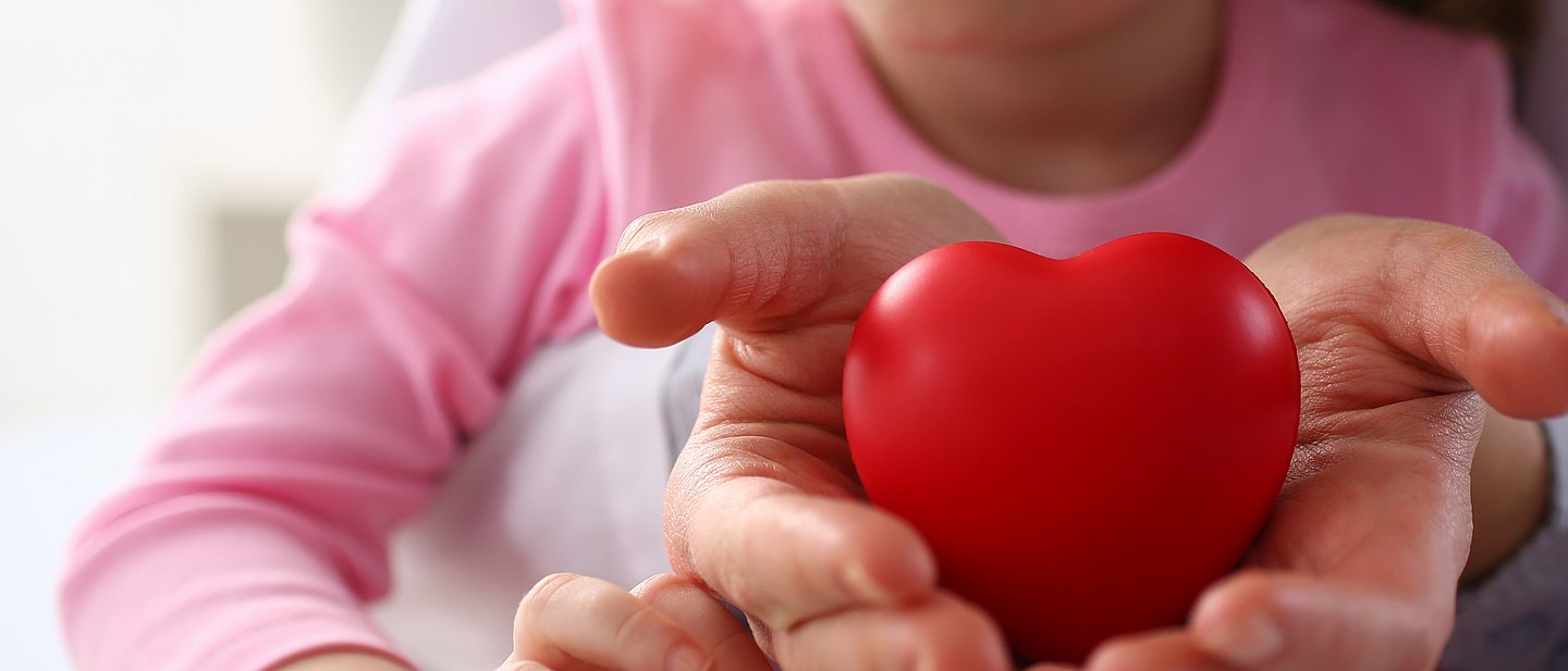zwei Hände halten ein rotes Herz, im Hintergrund ist unscharf der Körper eines Kinds auf einem Schoß sitzend zu sehen