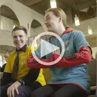 Zwei junge Menschen sitzen nebeneinander in einer Kirche und lächeln. Über dem Bild liegt ein Abspiel-Symbol, das auf ein Video hinweist.