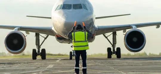 Ein Flughafenmitarbeiter in Warnweste weist auf dem Rollfeld ein großes Passagierflugzeug ein. Das Flugzeug steht frontal vor ihm, während der Mitarbeiter mit leuchtenden Signalkellen Anweisungen gibt.