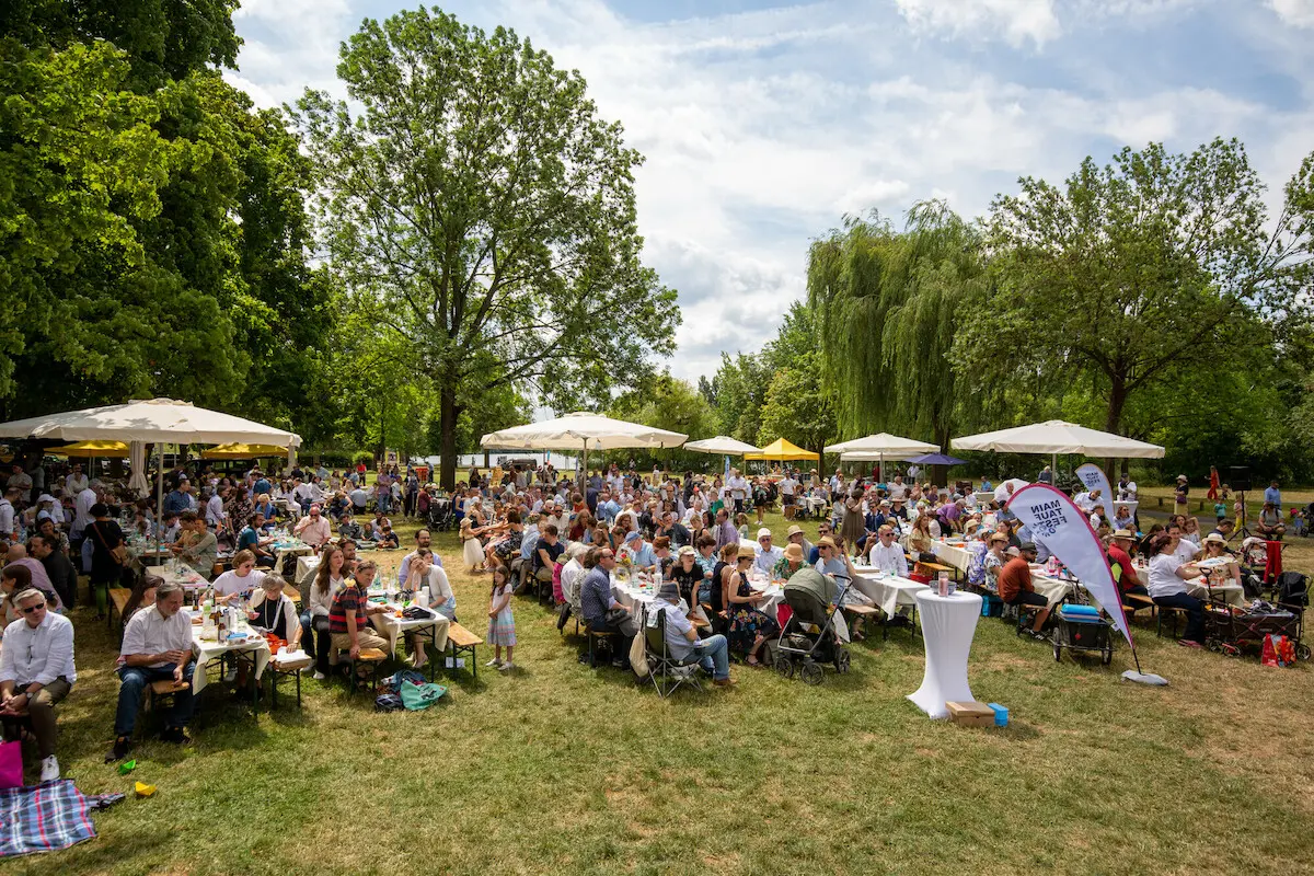 Hunderte Leute sitzen bei Sonnenschein unter Sonnenschirmen auf Bierbänken und feiern das "Main Tauffest 2022" am Mainufer in Offenbach.