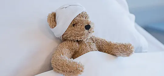 Ein Teddybär liegt im Krankenhausbett und trägt einen Verband am Kopf. Die Szene wirkt ruhig und kindgerecht