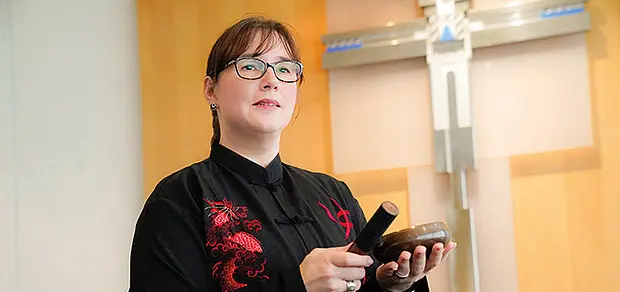 Eine Frau mit Brille steht in einem Andachtsraum vor einem Kreuz und hält eine Klangschale sowie einen Schlägel in den Händen. Sie trägt dunkle Kleidung mit roten Verzierungen und blickt aufmerksam nach vorne.